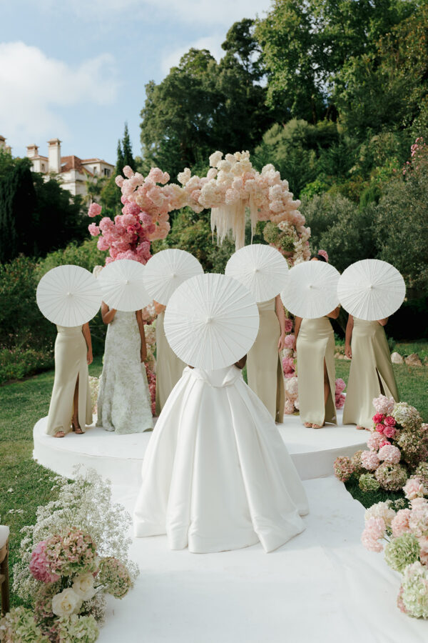 Bride and bridesmaids hiding their faces behind white parasols beneath a pink floral arch at Quinta da Bella Vista, Sintra.